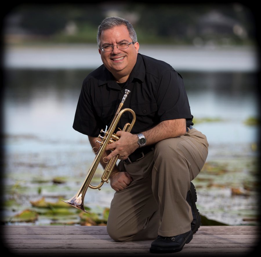Allen J. Venezio crouching by a lake with his trumpet